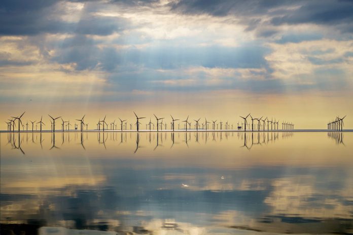 Offshore wind turbines silhouetted against a dramatic cloudscape, reflected in mirror-calm sea water