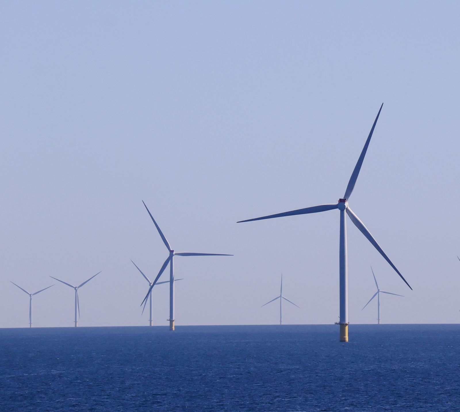 Row of offshore wind turbines at sea, blades turning against a pale sky